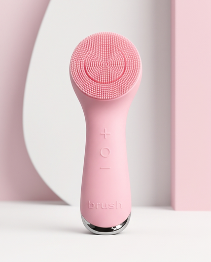A pink electric facial cleansing brush with a silicone brush head and a sleek handle, standing upright on a white surface with a minimalist pink and white background.